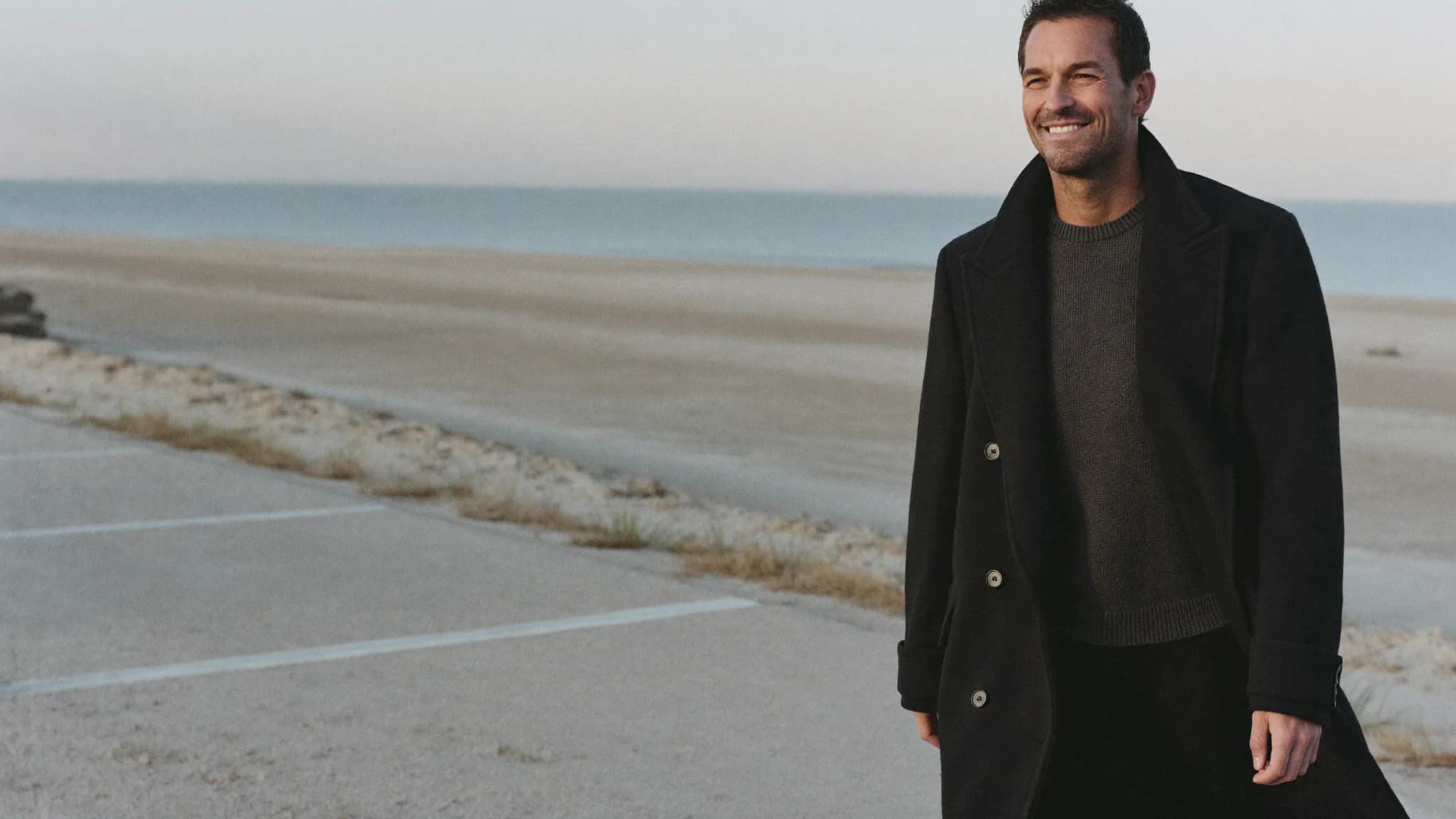 A man in a black coat walking along the shore
